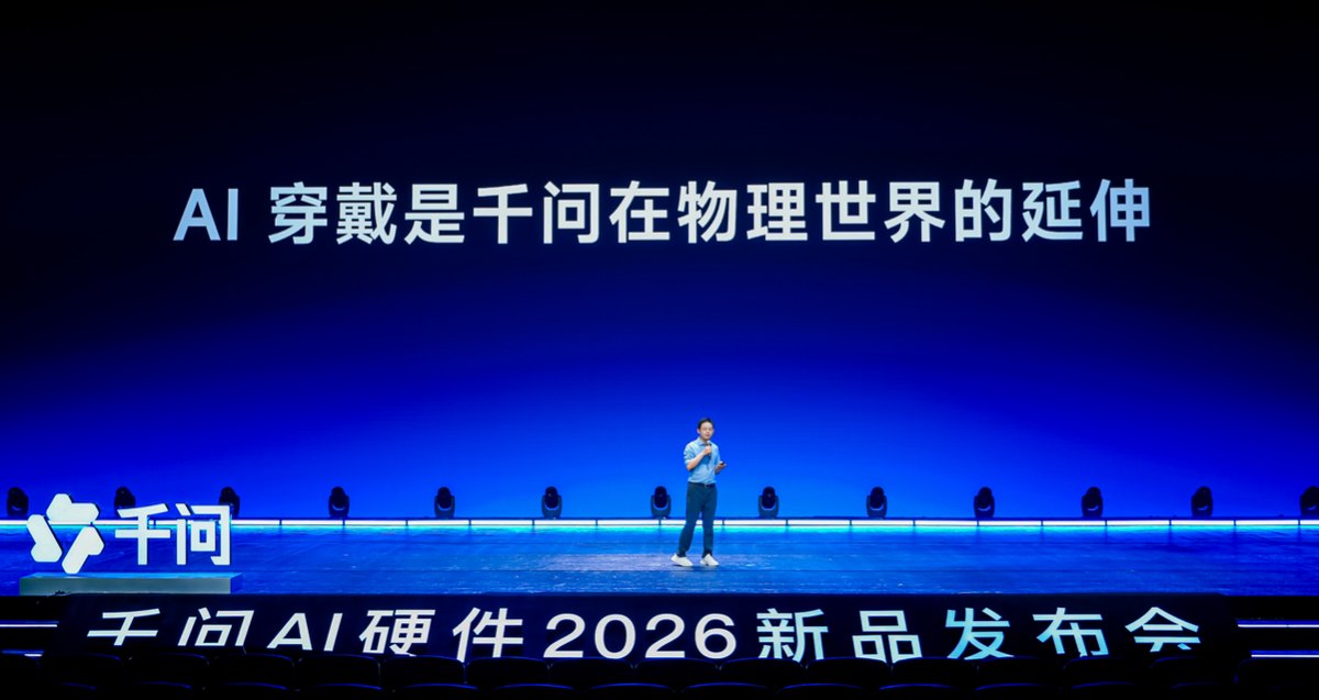 千问 AI 硬件 2026 新品发布会现场，讲者站在大屏前，屏幕写着 AI 穿戴是千问在物理世界的延伸